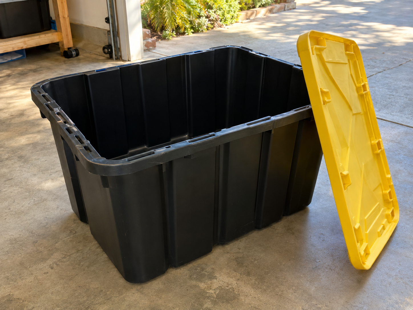 Open black moving bin with a yellow lid leaning beside it in a garage